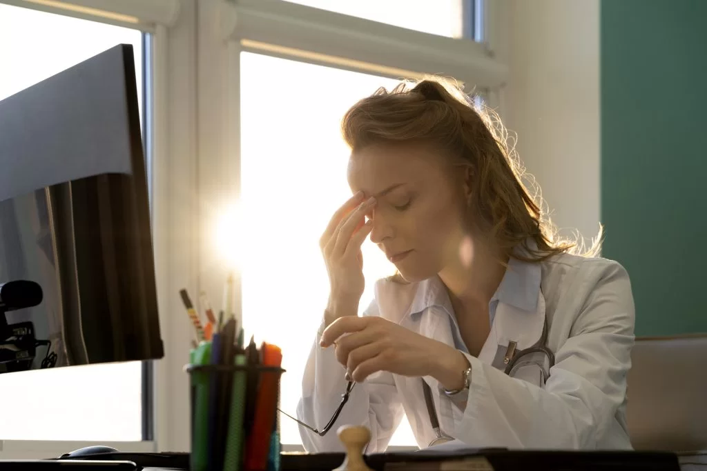 Clinician in a white coat at her desk pressing her fingers to her forehead with eyes closed