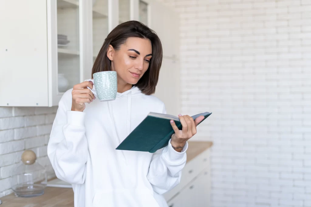 Young woman warm white hoodie home kitchen reads book