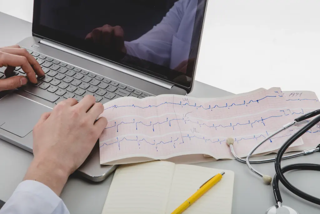 Doctor typing on a laptop beside an electrocardiogram (ECG) printout and stethoscope.