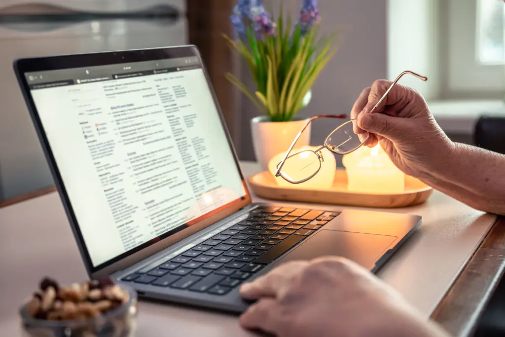Older hands resting near a laptop screen, with eyeglasses visible in the foreground; close-up view.