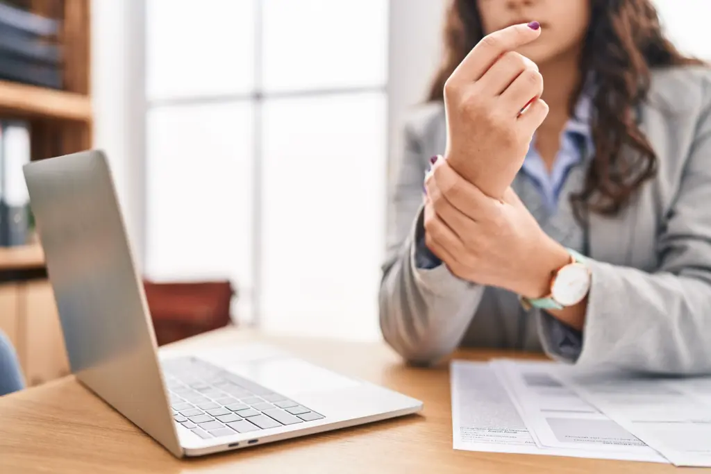 Woman experiencing wrist pain while working at a desk in an office, holding her sore wrist with one hand.